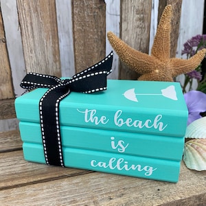 May include: A stack of three teal blue wooden blocks with white text that reads "The beach is calling". The top block has a white silhouette of an umbrella. The blocks are tied together with a black ribbon with white stitching.