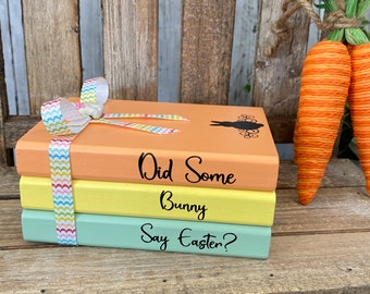 May include: Three stacked wooden blocks painted in pastel colours, orange, yellow, and green. Each block has a different Easter-themed phrase written in black: "Did Some Bunny Say Easter?"