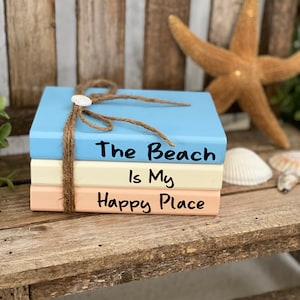 May include: Three stacked wooden blocks painted in shades of blue, cream, and peach. The top block reads "The Beach is My Happy Place" in black lettering.