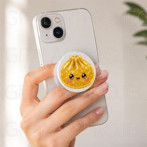 May include: A white smartphone featuring a circular phone grip attached to the back. The grip displays a gold-colored, glittery illustration of a steamed dumpling with a smiling face, large black eyes, and small pink cheeks on a white background.