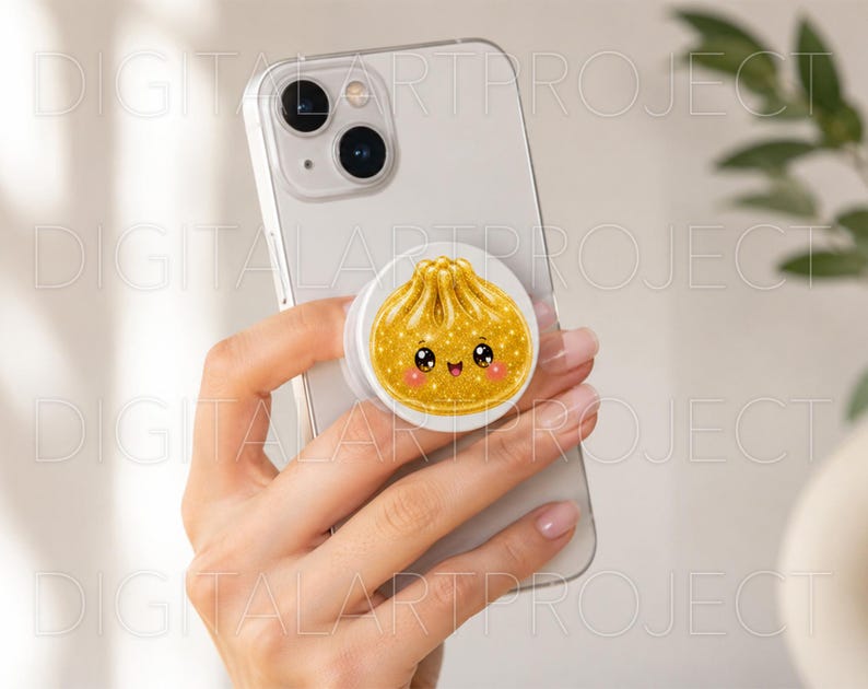 May include: A white smartphone featuring a circular phone grip attached to the back. The grip displays a gold-colored, glittery illustration of a steamed dumpling with a smiling face, large black eyes, and small pink cheeks on a white background.
