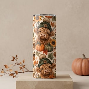 May include: A cylindrical tumbler featuring a repeating pattern of brown Highland cows wearing green knit beanies with pom-poms. The design includes orange and green pumpkins, yellow sunflowers, autumn leaves, and coffee cups on a cream background.