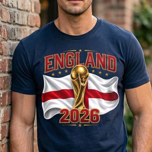 May include: Navy blue t-shirt featuring the word "ENGLAND" in red and gold lettering, with a gold trophy and the England flag. The number "2026" is displayed in red and gold below the flag.