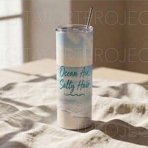Puede incluir: Un vaso blanco y azul con una pajita de acero inoxidable. El vaso tiene las palabras "Ocean Air, Salty Hair" en escritura turquesa. El dise&ntilde;o presenta una escena de playa con olas y un cielo azul claro.