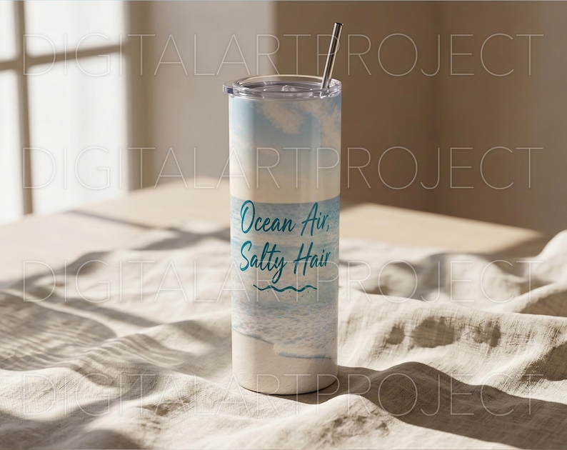 Puede incluir: Un vaso blanco y azul con una pajita de acero inoxidable. El vaso tiene las palabras "Ocean Air, Salty Hair" en escritura turquesa. El dise&ntilde;o presenta una escena de playa con olas y un cielo azul claro.