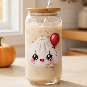 May include: A clear glass tumbler with a bamboo lid and a glass straw, filled with a beige iced beverage. The glass features a graphic of a white dumpling with a smiling face, red clown makeup, a red balloon, and a small paper boat.