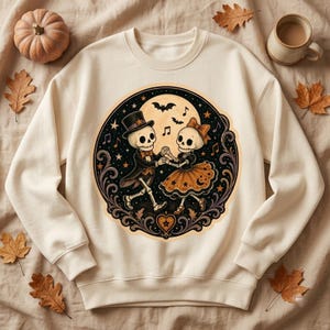 May include: A cream-colored crewneck sweatshirt featuring a circular graphic of two skeletons dancing. One skeleton wears a top hat and tuxedo, while the other wears a bow and a skirt. The design includes a full moon, stars, bats, and musical notes.