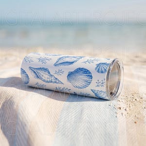 Puede incluir: Un vaso blanco y azul con estampado de conchas marinas y tapa transparente descansa sobre una toalla de playa a rayas. El vaso presenta varias ilustraciones de conchas marinas en tonos azules, creando un tema costero. El fondo muestra el oc&eacute;ano y el cielo.