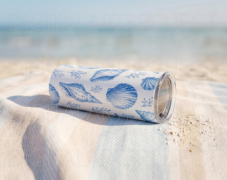 Puede incluir: Un vaso blanco y azul con estampado de conchas marinas y tapa transparente descansa sobre una toalla de playa a rayas. El vaso presenta varias ilustraciones de conchas marinas en tonos azules, creando un tema costero. El fondo muestra el oc&eacute;ano y el cielo.