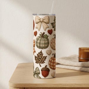 May include: A cylindrical tumbler with a straw featuring a cream-colored background decorated with a repeating pattern of beige bows, green plaid pumpkins, orange pumpkins, leopard print maple leaves, brown acorns, and small red and white hearts.