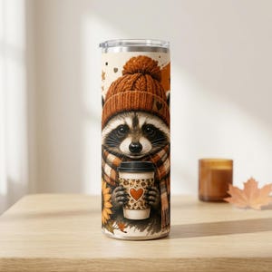 May include: A cylindrical tumbler featuring a graphic of a raccoon wearing a brown knit beanie and a plaid scarf. The raccoon holds a coffee cup with a leopard print pattern and a heart icon. The design includes sunflowers and autumn leaves.