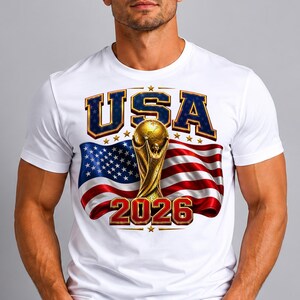 May include: White t-shirt featuring a graphic design with the text "USA" in blue and gold, a golden trophy, and the number "2026" in red. The design includes the American flag in the background.