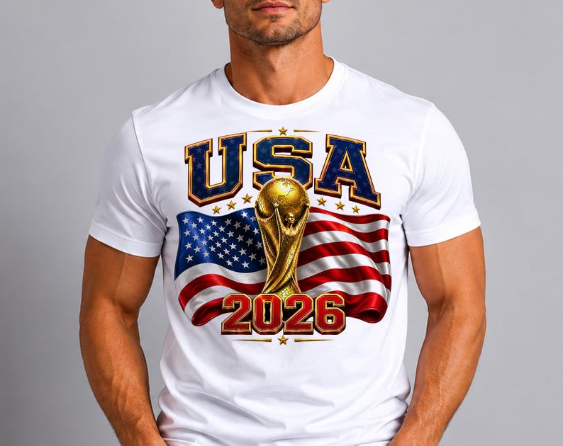 May include: White t-shirt featuring a graphic design with the text "USA" in blue and gold, a golden trophy, and the number "2026" in red. The design includes the American flag in the background.
