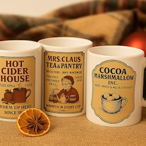 May include: Three white ceramic mugs with festive labels. One reads "Hot Cider House" with apple and clove illustrations. Another says "Mrs. Claus Tea & Pantry." The third mug is labeled "Cocoa Marshmallow Inc."