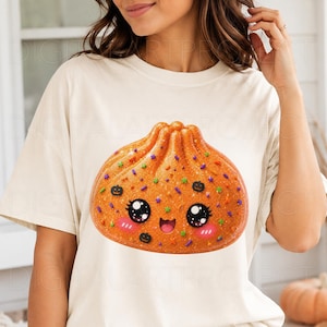May include: A cream-colored short-sleeve t-shirt featuring a large, orange, glittery dumpling graphic. The dumpling has a smiling face with large black eyes, pink cheeks, and is decorated with small green star and black pumpkin sprinkles.