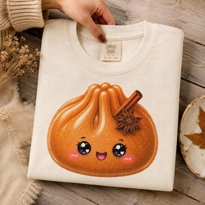 May include: A cream-colored crewneck sweatshirt featuring a graphic of a golden-brown dumpling with a smiling face, pink cheeks, and large eyes. A cinnamon stick and a star anise are placed on top of the dumpling illustration. The inner neck label reads COMFORT COLORS.