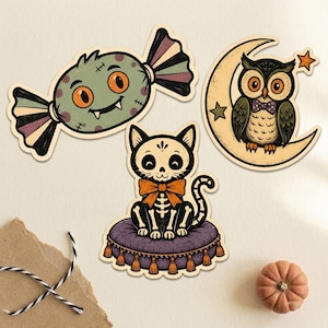 May include: Three die-cut stickers featuring a green monster candy with orange eyes, a black cat skeleton wearing an orange bow tie sitting on a purple tasseled cushion, and a brown owl perched on a yellow crescent moon with two stars.