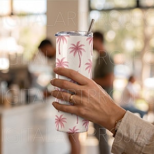 Puede incluir: Un vaso de acero inoxidable blanco con un estampado de palmeras rosas. El vaso tiene una pajita plateada y una tapa. El dise&ntilde;o presenta palmeras rosas repetidas sobre un fondo blanco. El vaso lo sostiene una persona.