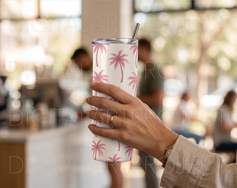 Puede incluir: Un vaso de acero inoxidable blanco con un estampado de palmeras rosas. El vaso tiene una pajita plateada y una tapa. El dise&ntilde;o presenta palmeras rosas repetidas sobre un fondo blanco. El vaso lo sostiene una persona.