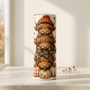May include: A cylindrical tumbler featuring a vertical stack of three fluffy Highland cows. The top cow wears an orange knit beanie, the middle cow has a plaid scarf, and the bottom cow wears a leopard print bow. Three pumpkins sit at the base.