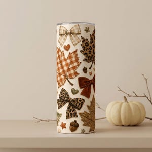 May include: A cylindrical tumbler featuring a cream-colored background decorated with a repeating pattern of brown and orange plaid maple leaves, leopard print maple leaves, solid brown bows, plaid bows, leopard print bows, acorns, and small hearts.