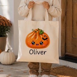 May include: A beige canvas tote bag featuring a graphic of a smiling orange pumpkin dumpling with a green leaf, a small black bat, and a crescent moon. The name Oliver is printed in black text below the pumpkin illustration.