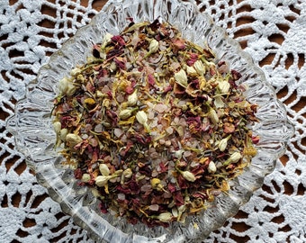 May include: A clear, decorative glass bowl filled with a mixture of dried flowers, buds, and salt crystals. The mix includes red, white, and yellow petals, with white jasmine buds. The bowl sits on a white, open-weave crocheted doily.