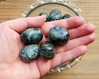 May include: A collection of six green and black tumbled stones with a swirling pattern. The stones are in a person's hand, which is holding them over a clear glass bowl.