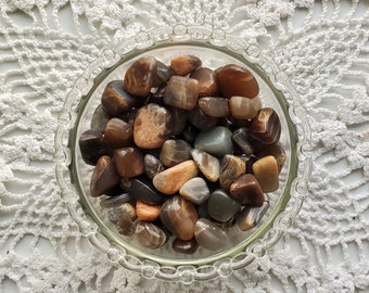 May include: A glass bowl filled with a variety of smooth, polished stones in shades of brown, gray, and orange. The stones are arranged in a random pattern and appear to be natural.