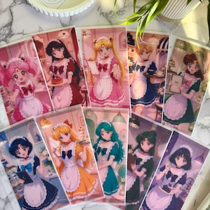 May include: A collection of nine rectangular anime-style character cards featuring figures in maid outfits with white aprons and lace headpieces. Each card displays a different character with unique hair colors and poses, holding items like trays, menus, or desserts.