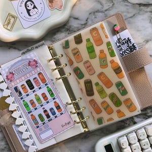 Op de afbeelding: Een beige planner met gouden accenten, open om decoratieve inzetstukken te tonen. E&eacute;n inzetstuk toont een frisdrankbespaartabel en een ander heeft een patroon van frisdrankblikjes en -flessen. Een rekenmachine en stickers zijn ook zichtbaar.