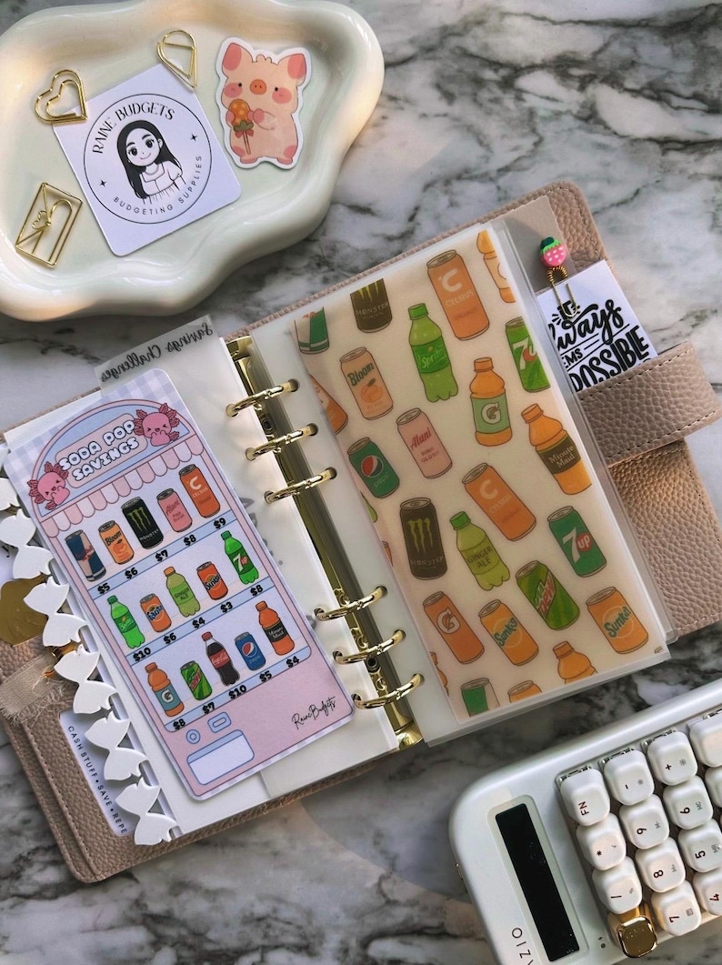Op de afbeelding: Een beige planner met gouden accenten, open om decoratieve inzetstukken te tonen. E&eacute;n inzetstuk toont een frisdrankbespaartabel en een ander heeft een patroon van frisdrankblikjes en -flessen. Een rekenmachine en stickers zijn ook zichtbaar.