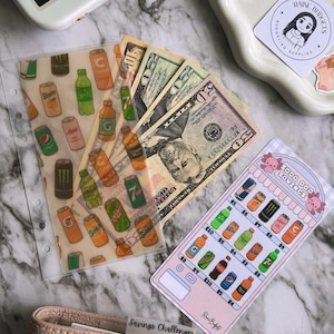 Op de afbeelding: Een doorzichtige plastic envelop met een frisdrankontwerp, bankbiljetten en een spaartrackerkaart. De kaart heeft een frisdrankthema met prijzen. Een beige portemonnee met de tekst "Savings Challenges" is ook zichtbaar.