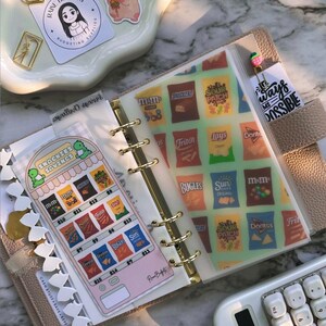 Op de afbeelding: Een beige planner met gouden ringen, open om decoratieve inzetstukken te laten zien. Een inzetstuk heeft een snack-thema-ontwerp met verschillende chipmerken. Een bladwijzer leest "Snackies Savings". Een rekenmachine en stickers zijn ook zichtbaar.