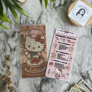 May include: A clear plastic sleeve featuring a Hello Kitty chef illustration with a strawberry shortcake. A recipe list for strawberry shortcake is next to it. A wooden lid with a sticker that says "RAINE BUDGETS BUDGETING SUPPLIES" is also in the image.