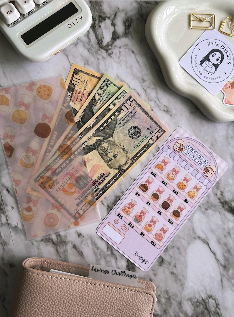 Op de afbeelding: Een bovenaanzicht toont Amerikaanse bankbiljetten naast een pastelroze spaartracker met gebakillustraties. Een doorzichtig zakje met een dessertpatroon en een lichtroze portemonnee zijn ook zichtbaar. Een witte rekenmachine staat op de achtergrond.