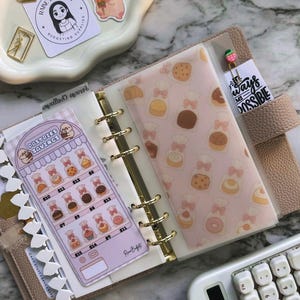 Op de afbeelding: Een beige planner met gouden ringen, open om decoratieve inzetstukken te laten zien. E&eacute;n inzetstuk heeft een gebakthema, terwijl een ander een spaartracker weergeeft. Een witte rekenmachine en decoratieve paperclips zijn ook zichtbaar.