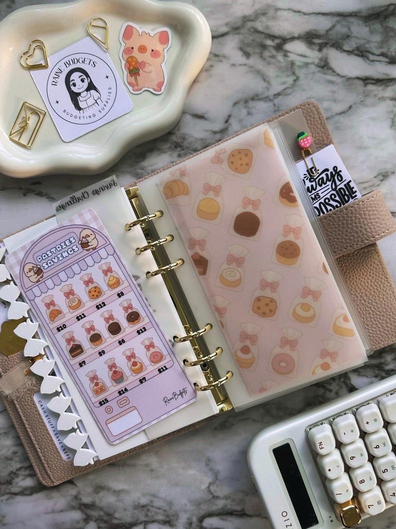Op de afbeelding: Een beige planner met gouden ringen, open om decoratieve inzetstukken te laten zien. E&eacute;n inzetstuk heeft een gebakthema, terwijl een ander een spaartracker weergeeft. Een witte rekenmachine en decoratieve paperclips zijn ook zichtbaar.
