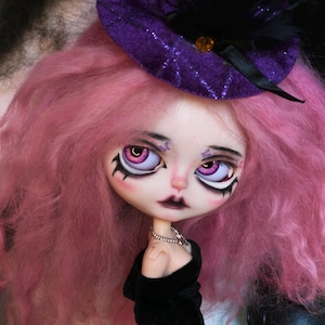 May include: A doll with long, wavy pink hair wearing a purple glittery hat adorned with black feathers and a small amber bead. The doll features large purple eyes, dark eyeliner, and small butterfly accents above the eyes, wearing a black dress.
