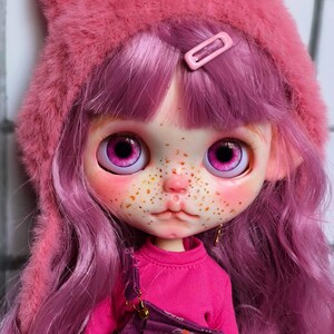 May include: A doll with a pink cat-ear hat, pink hair, and a pink outfit. The doll has large, expressive eyes and freckles. The doll is wearing a pink shirt and purple overalls.
