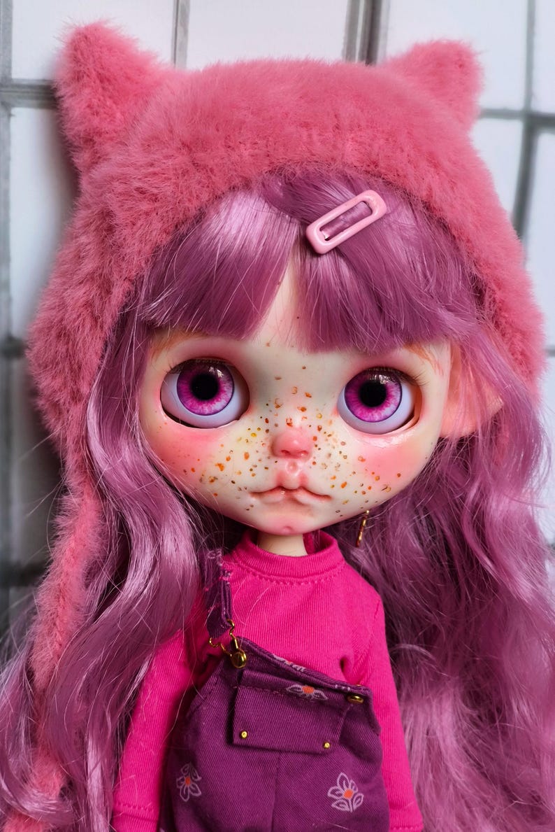 May include: A doll with a pink cat-ear hat, pink hair, and a pink outfit. The doll has large, expressive eyes and freckles. The doll is wearing a pink shirt and purple overalls.