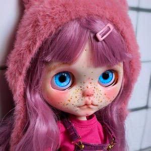 May include: A doll with bright blue eyes, freckles, and pink hair. The doll wears a pink fuzzy hat with cat ears, a pink shirt, and purple overalls. A pink barrette secures the hair.