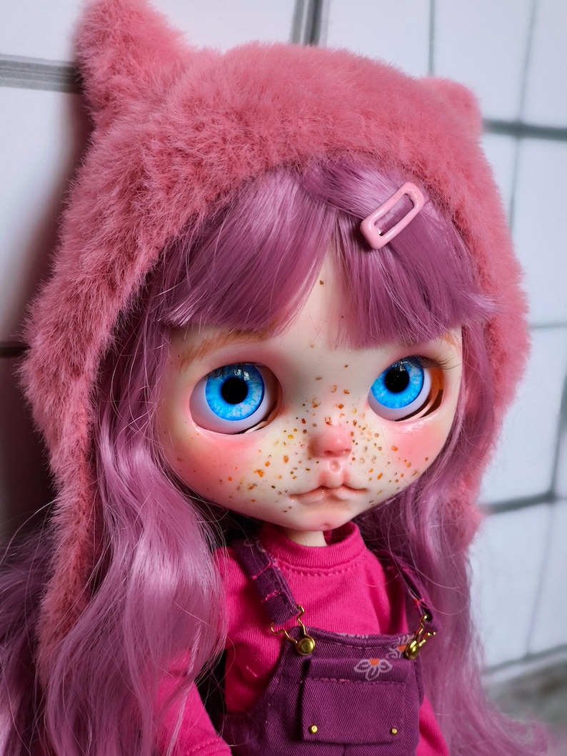 May include: A doll with bright blue eyes, freckles, and pink hair. The doll wears a pink fuzzy hat with cat ears, a pink shirt, and purple overalls. A pink barrette secures the hair.