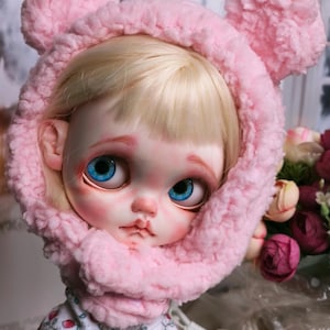 May include: A doll with blonde hair and blue eyes wearing a pink, fuzzy hood with bear ears. The doll is dressed in a white dress with a red floral pattern. The doll's face has a delicate, painted appearance.