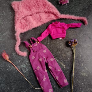 May include: A doll clothing set featuring a pink fuzzy hat, a magenta turtleneck sweater, and magenta overalls with a floral pattern. The set is arranged on a dark surface with dried flowers.