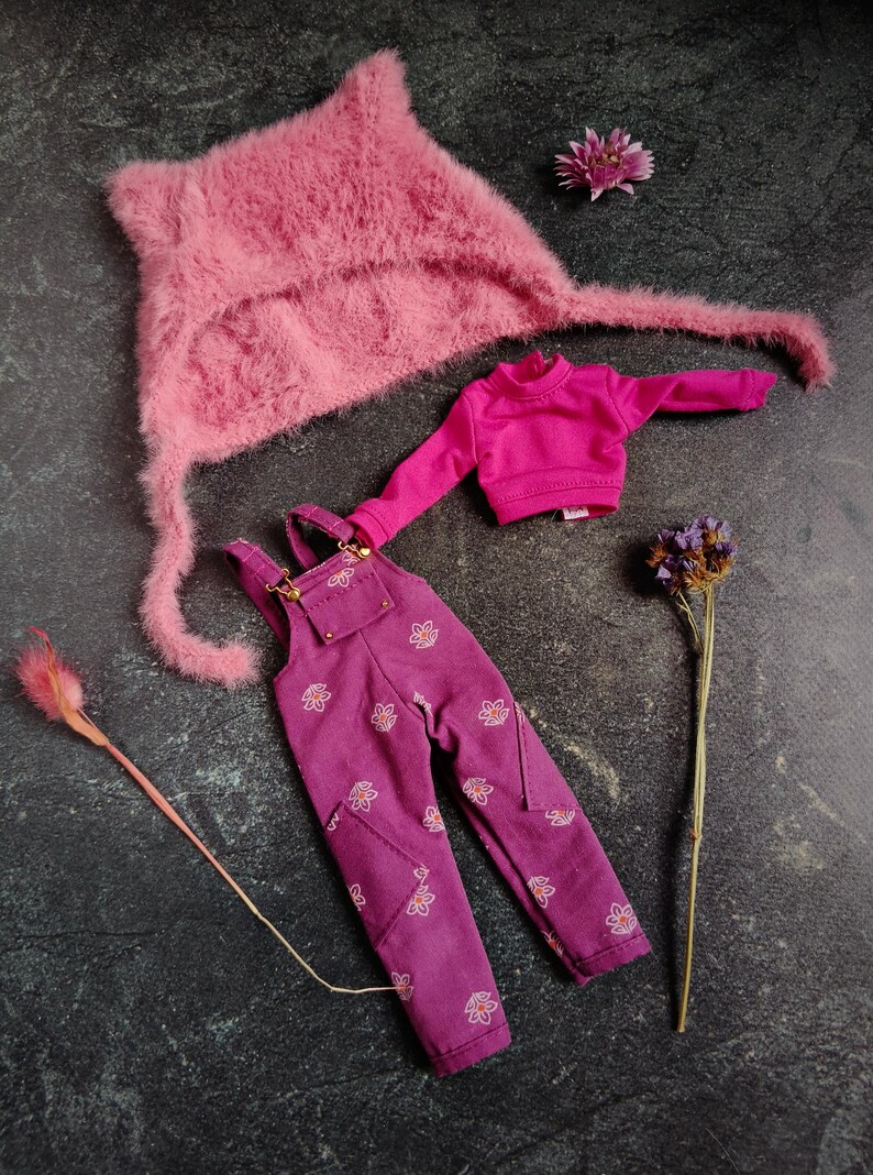 May include: A doll clothing set featuring a pink fuzzy hat, a magenta turtleneck sweater, and magenta overalls with a floral pattern. The set is arranged on a dark surface with dried flowers.