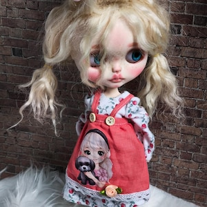 May include: A handmade doll with blonde hair styled in pigtails and a pink bow. The doll wears a red dress with a graphic of a girl and a dog. The doll has blue eyes and is standing in front of a brick wall.