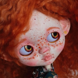 May include: A handmade doll with red curly hair and freckles. The doll has large, expressive eyes with yellow and red irises. It wears a dark green floral dress and a small gold earring. The doll's skin is pale with rosy cheeks and lips.