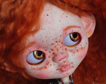 Può includere: Una bambola fatta a mano con capelli rossi ricci e lentiggini. La bambola ha grandi occhi con iridi gialle e rosse. Indossa un vestito floreale verde scuro e un piccolo orecchino d'oro. La pelle della bambola è pallida con guance e labbra rosee.