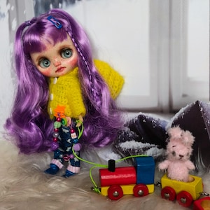 May include: A purple haired doll with a yellow sweater and blue pants is playing with a toy train and a pink teddy bear. The train is red, yellow, and blue with a black chimney. The teddy bear is sitting in a yellow wooden car.